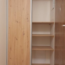 Xeraco Design - Wnętrze przedpokoju z częściowo otwartą szafą wnękową z przesuwnymi drzwiami o wzorze drewna, widoczne półki i fragment drzwi wejściowych.