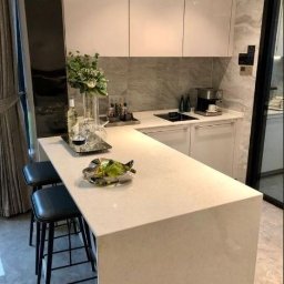 Xeraco Design - Nowoczesna kuchnia z białymi szafkami bez uchwytów, marmurowym blatem i wyspą z dwoma czarnymi hokerami, dekorowana kwiatami i owocami, z ekspressem do kawy w tle.