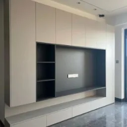 Nowoczesna zabudowa meblowa w salonie: beżowe szafki wiszące i dolne szuflady z ciemnoszarą wnęką na telewizor i półkami, marmurowa podłoga w odcieniach szarości.