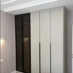 Nowoczesna szafa wnękowa z białymi frontami i czarnymi, długimi uchwytami, z jedną częścią przeszkloną z ciemnego szkła, w jasnym pomieszczeniu z listwą przysufitową.