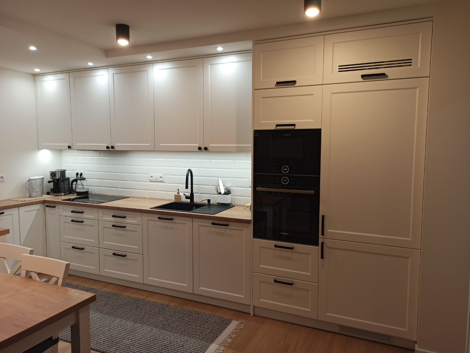 Nowoczesna kuchnia z białymi szafkami na wymiar, czarnym zlewem i armaturą, drewnianym blatem i wbudowanym piekarnikiem Bosch, oświetlona punktowymi lampami sufitowymi.