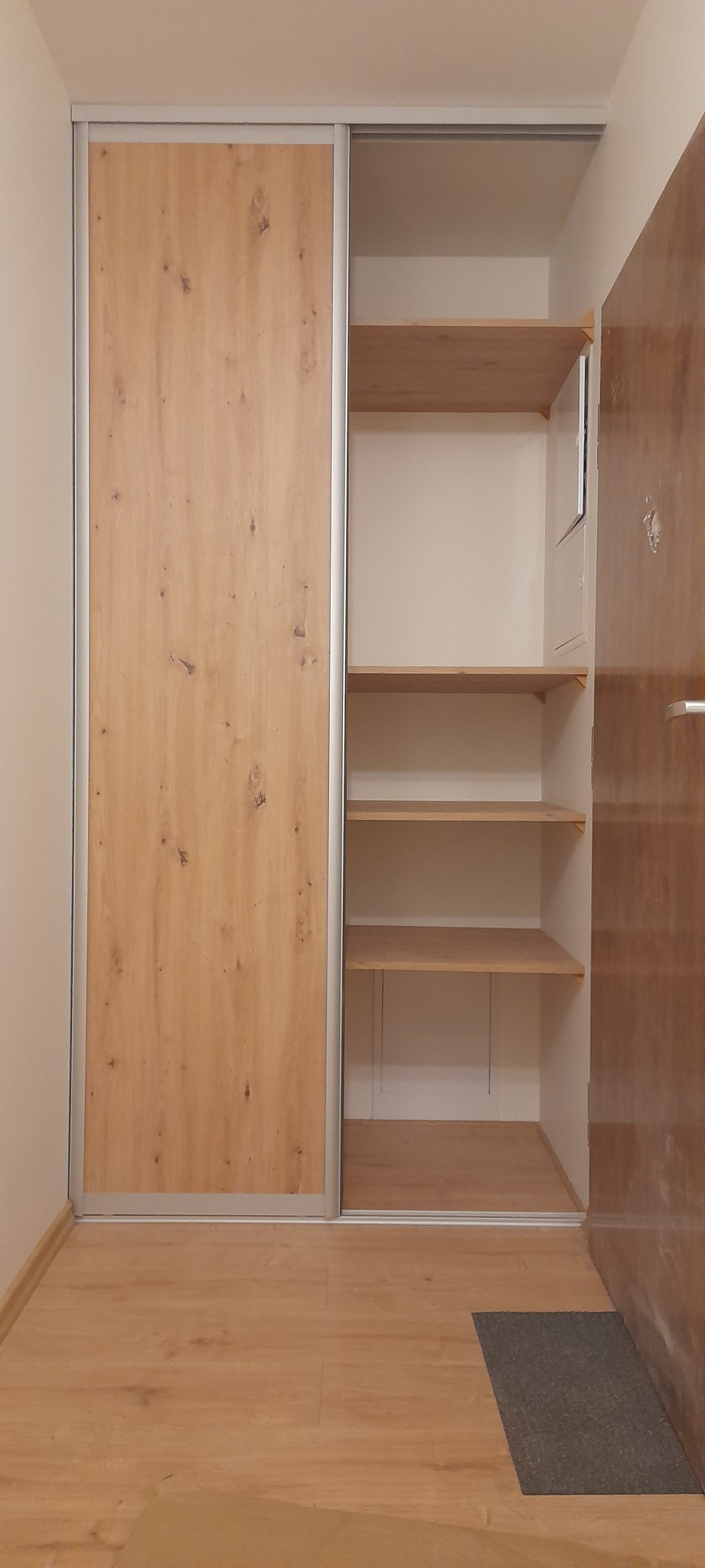 Wnętrze przedpokoju z częściowo otwartą szafą wnękową z przesuwnymi drzwiami o wzorze drewna, widoczne półki i fragment drzwi wejściowych.