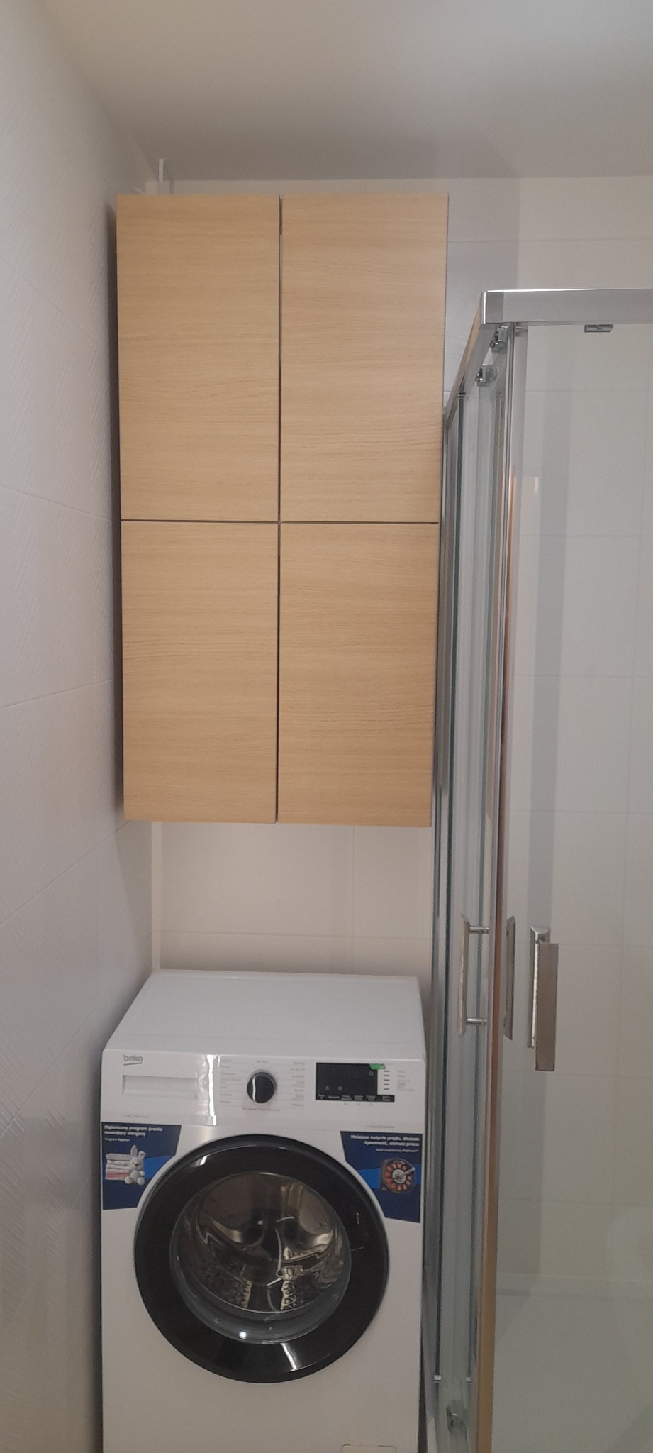 Jasna, drewnopodobna szafka wisząca nad białą pralką w łazience z kabiną prysznicową.