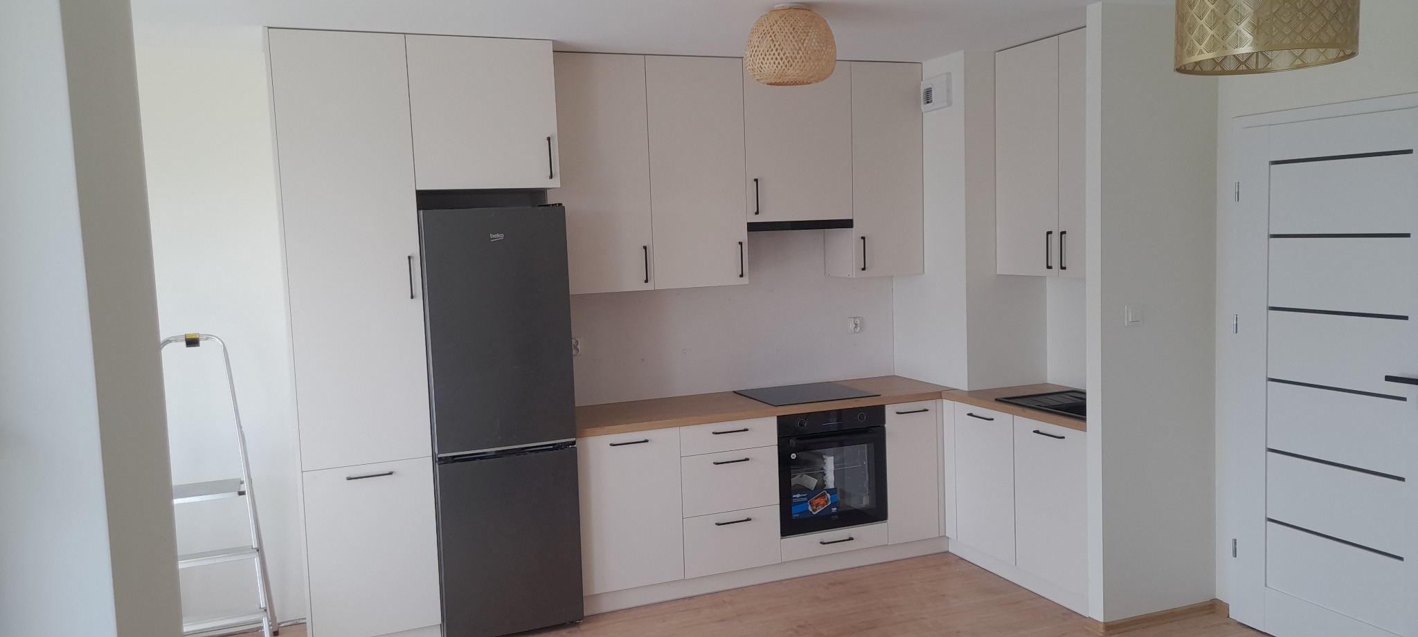 Jasna kuchnia z białymi szafkami z czarnymi uchwytami, szarą lodówką Beko i drewnianym blatem, widoczna drabina aluminiowa w rogu.