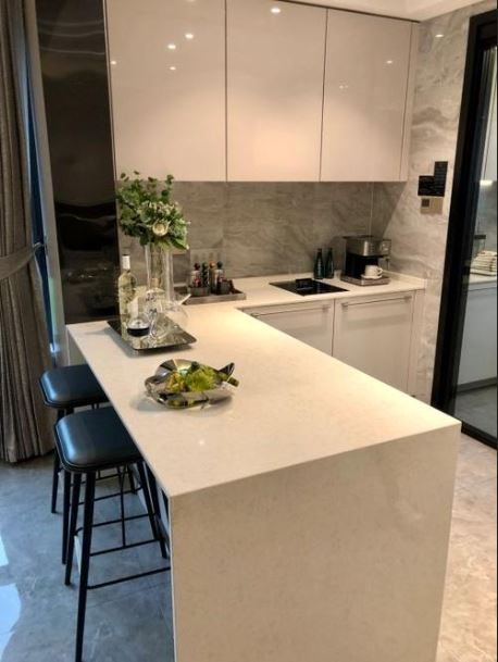 Nowoczesna kuchnia z białymi szafkami bez uchwytów, marmurowym blatem i wyspą z dwoma czarnymi hokerami, dekorowana kwiatami i owocami, z ekspressem do kawy w tle.