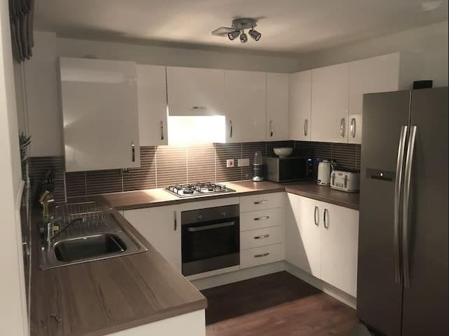 Nowoczesna kuchnia z białymi, błyszczącymi szafkami, brązowym blatem i płytkami, srebrną lodówką oraz oświetleniem punktowym.