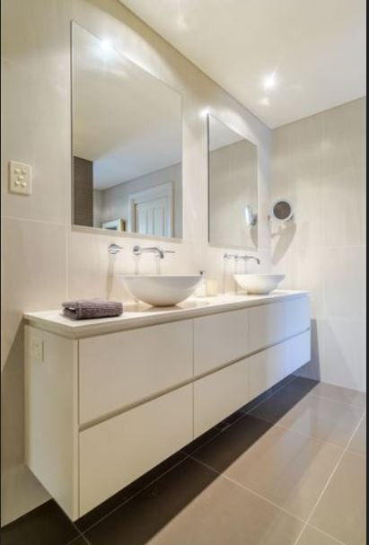 Nowoczesna łazienka z podwójną umywalką nablatową, długą szafką z białymi frontami na wysoki połysk i dwoma dużymi lustrami na ścianie wyłożonej jasnymi płytkami.