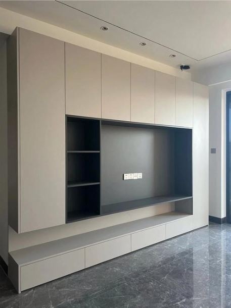 Nowoczesna zabudowa meblowa w salonie: beżowe szafki wiszące i dolne szuflady z ciemnoszarą wnęką na telewizor i półkami, marmurowa podłoga w odcieniach szarości.