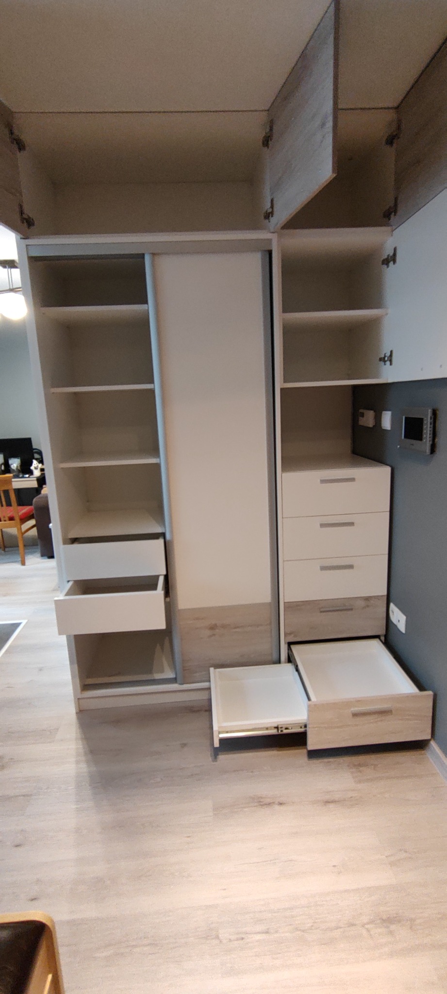 Biała szafa z przesuwnymi drzwiami i otwartymi szafkami, widoczne wysuwane szuflady, wnętrze w odcieniach bieli i drewna, zamontowana na ścianie, perspektywa od dołu ukazująca pełną wysokość.