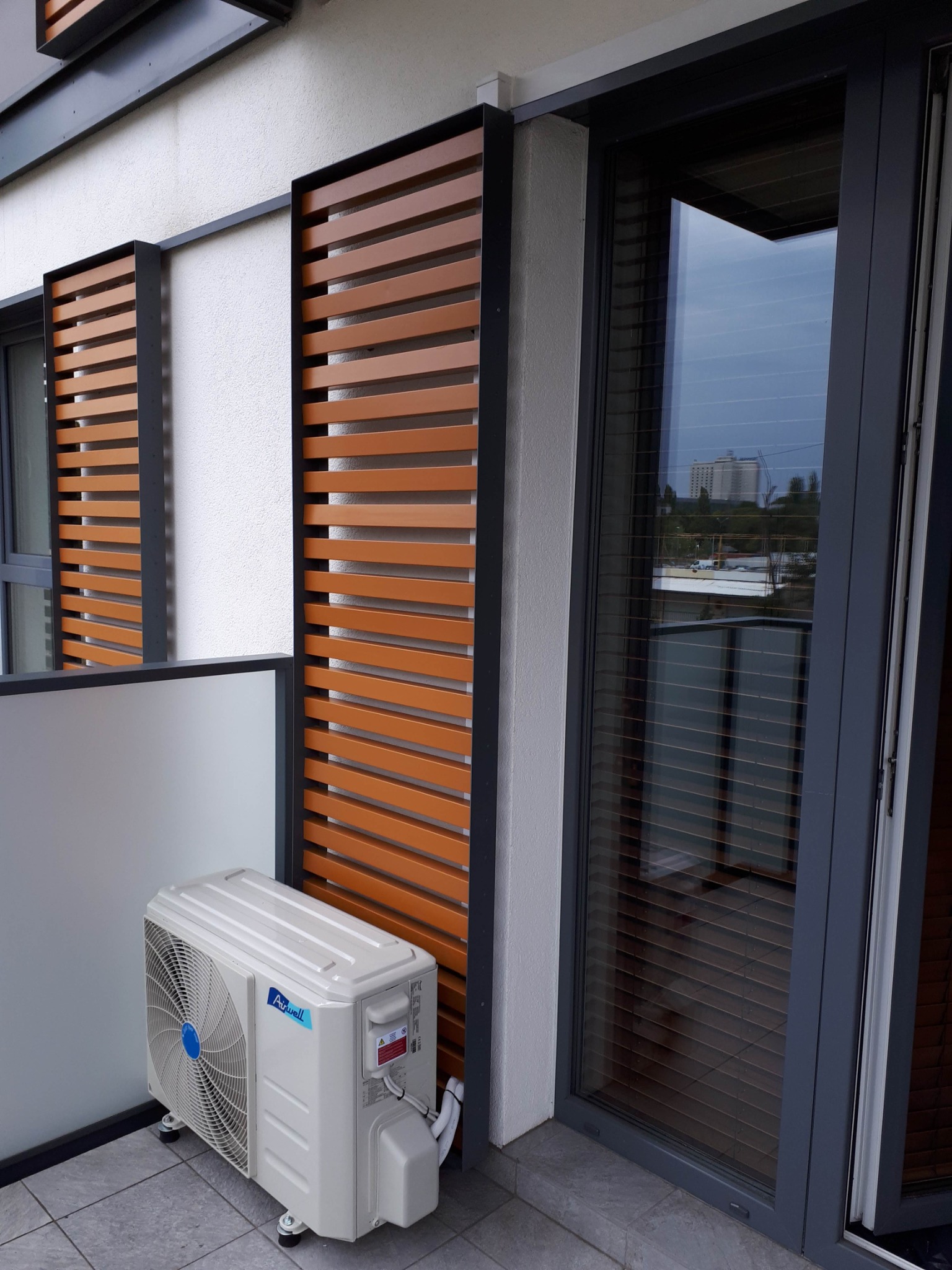 Biały klimatyzator zewnętrzny Airwell na balkonie z drewnianymi żaluzjami i szarymi ramami okiennymi, widok na miasto w odbiciu szyby.