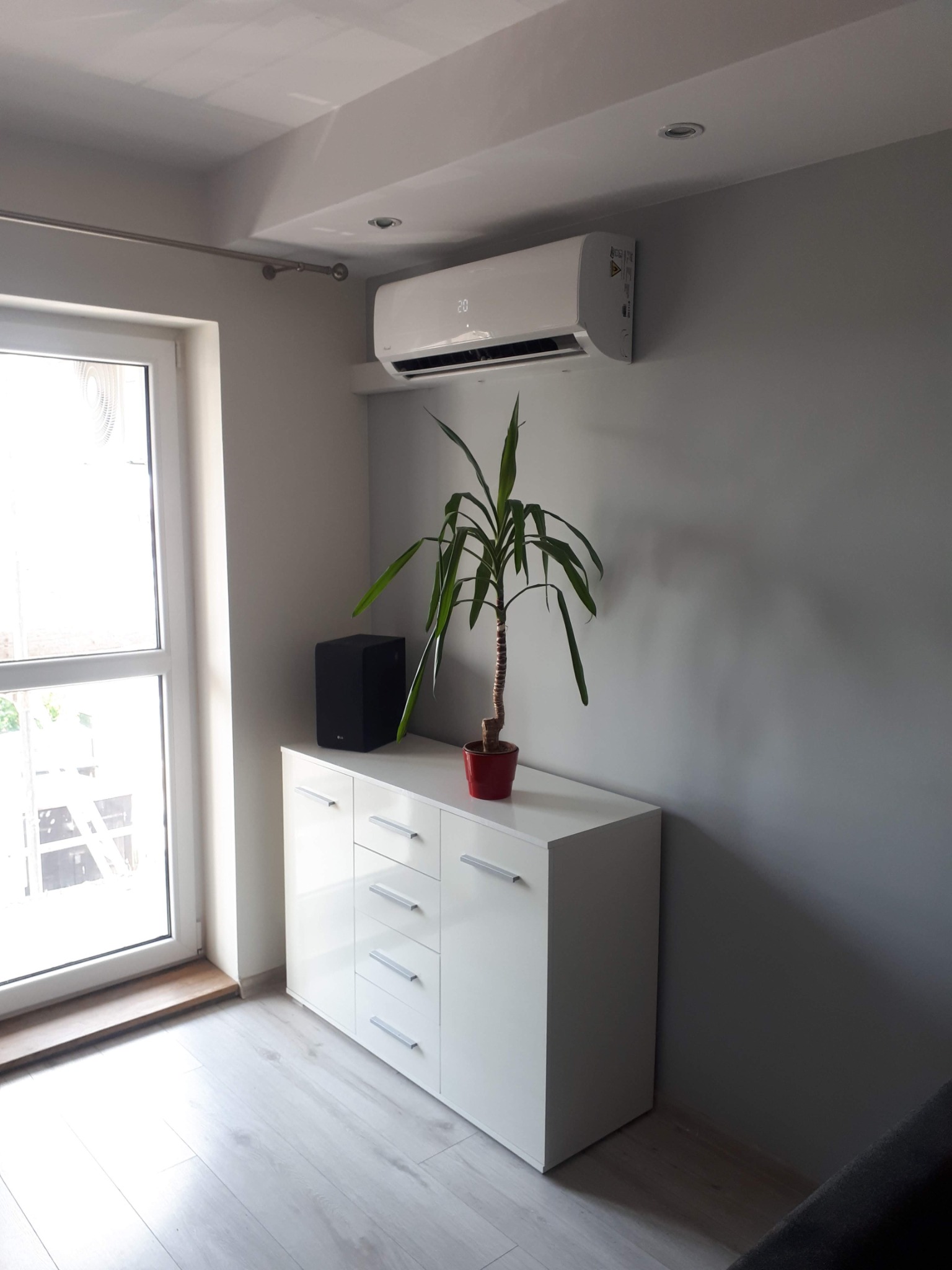 Biały klimatyzator ścienny z wyświetlaczem temperatury 20 stopni Celsjusza, zamontowany nad białą komodą z doniczką i głośnikiem, w jasnym, minimalistycznym wnętrzu.