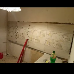 Ściana w trakcie wykańczania z ułożonymi płytkami z motywem liści, poziomica i krzyżyki dystansowe widoczne, butelka napoju na blacie.