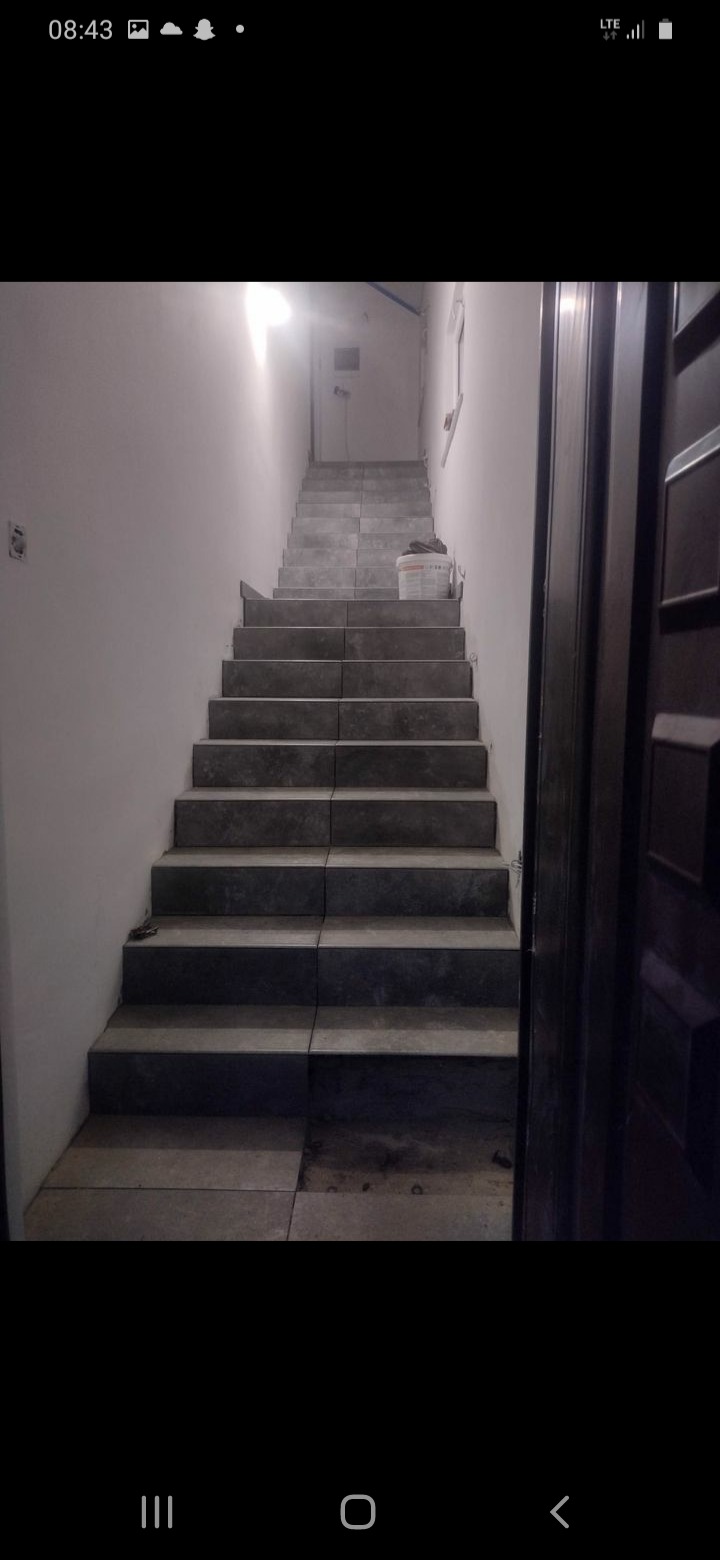 Schody wyłożone szarymi płytkami, widoczne niedokończone fragmenty na dole, wiadro z zaprawą na górze, surowe ściany w tle.