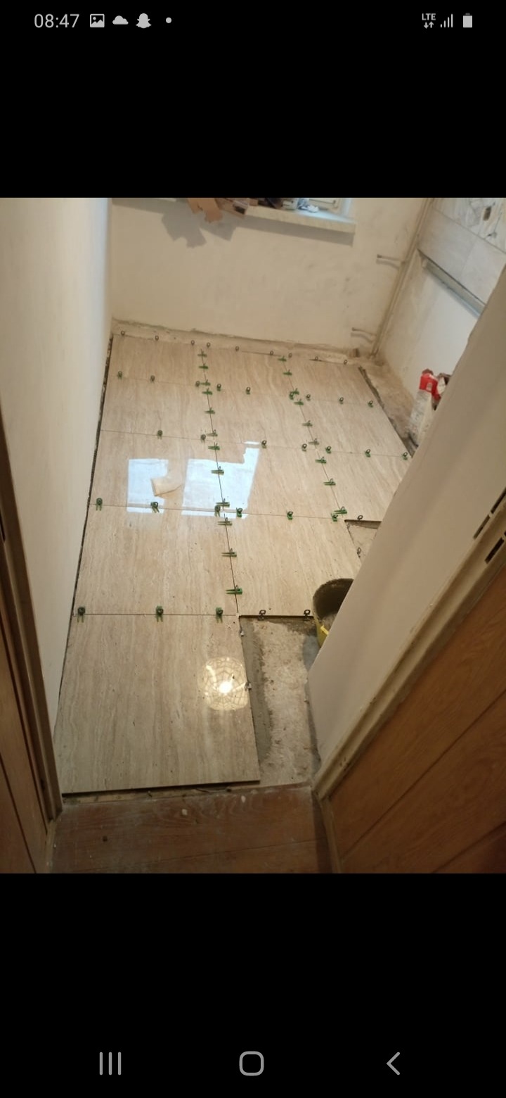 Beżowe płytki podłogowe, częściowo ułożone w łazience, z widocznymi krzyżykami dystansowymi i fragmentem nieukończonej podłogi.