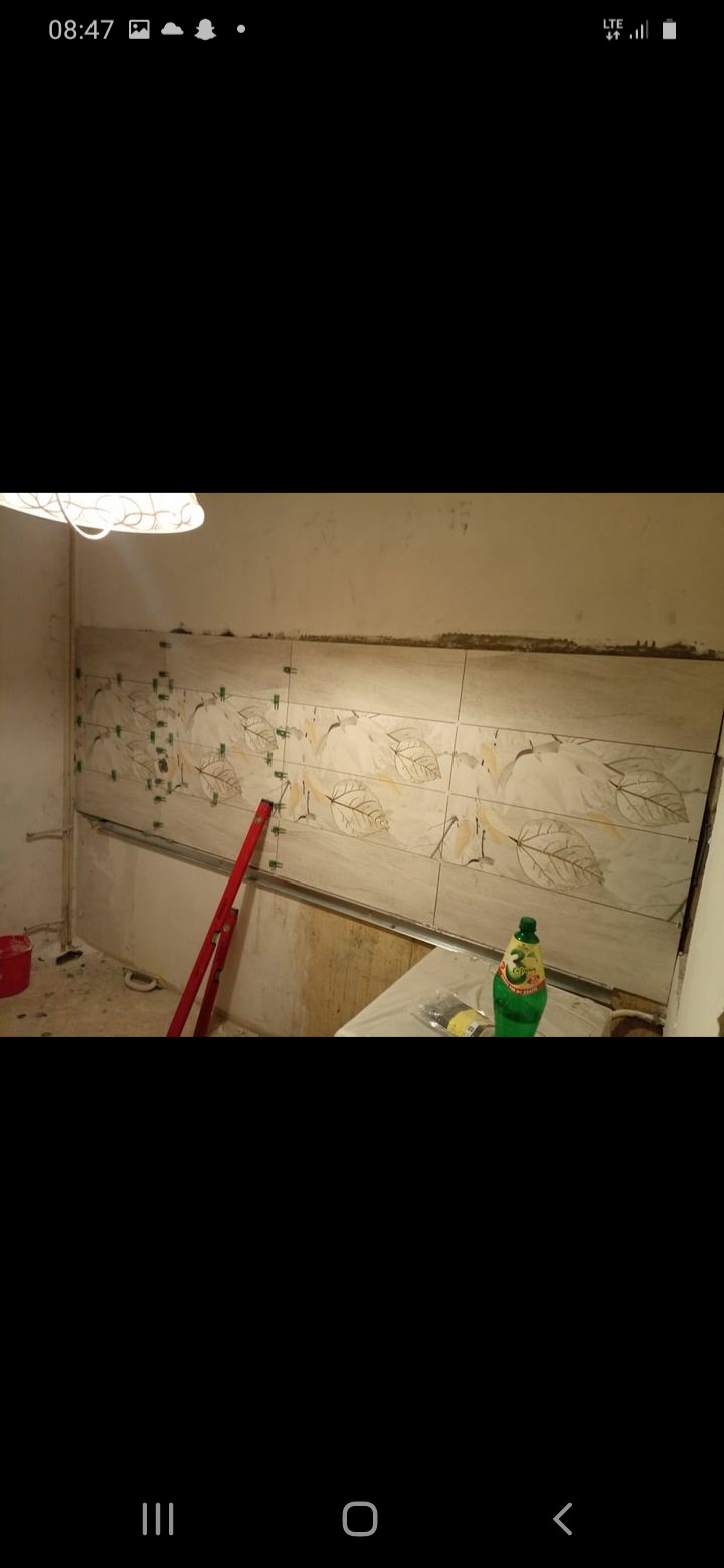 Ściana w trakcie wykańczania z ułożonymi płytkami z motywem liści, poziomica i krzyżyki dystansowe widoczne, butelka napoju na blacie.