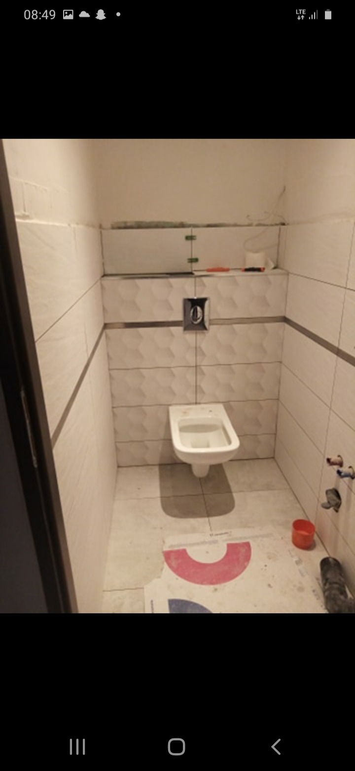 Montaż podwieszanej toalety w łazience z częściowo położonymi białymi płytkami z dekoracyjnym pasem, widoczne materiały budowlane i akcesoria.