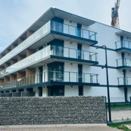 Nowoczesny, biały budynek wielorodzinny z przeszklonymi balkonami i gabionowym ogrodzeniem, w tle żurawie budowlane. Elewacja wykończona, stan deweloperski.