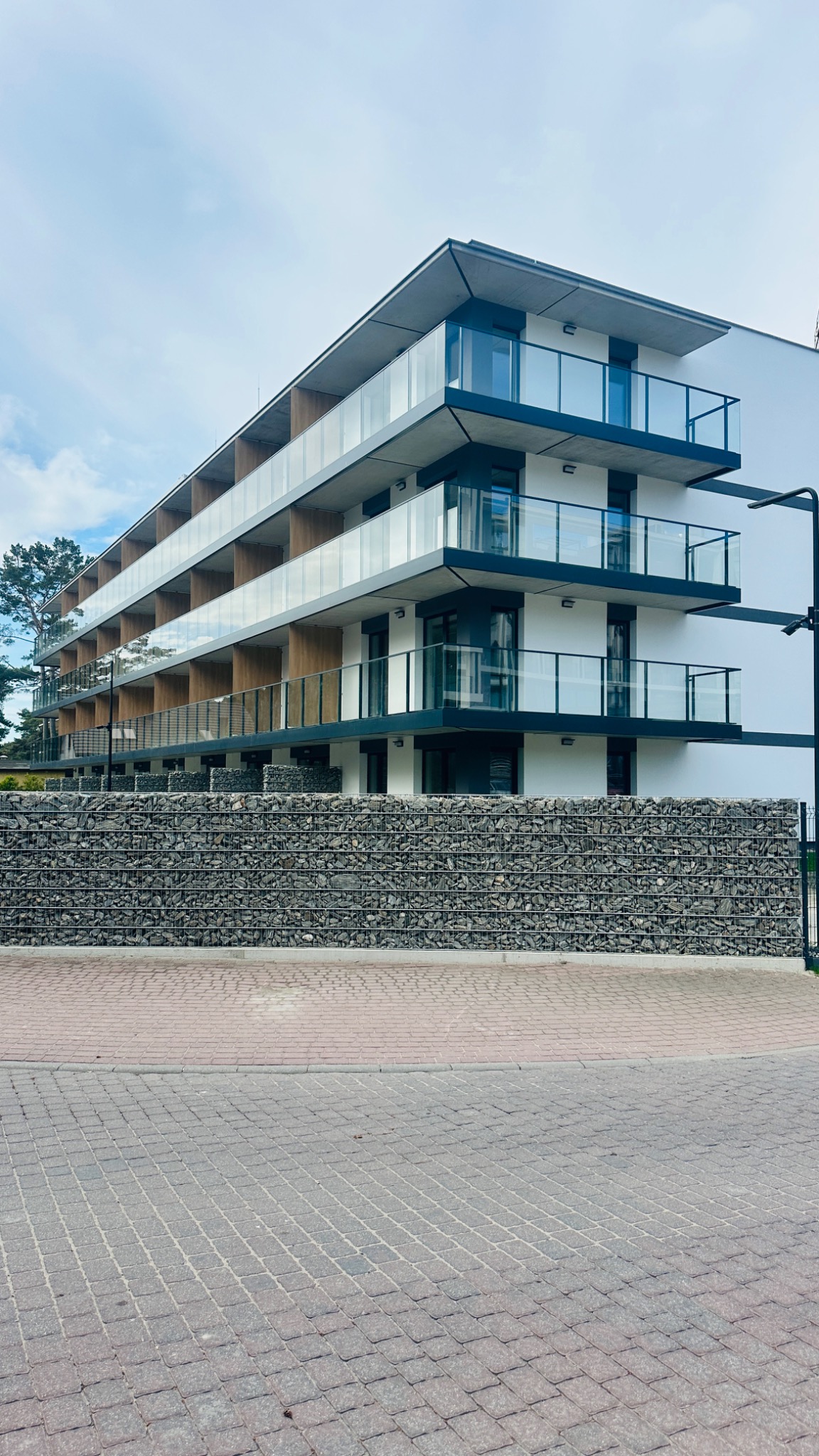 Nowoczesny budynek wielopiętrowy z białymi elewacjami i szklanymi balkonami, otoczony gabionami z kamienia. Widok z poziomu brukowanej ulicy.