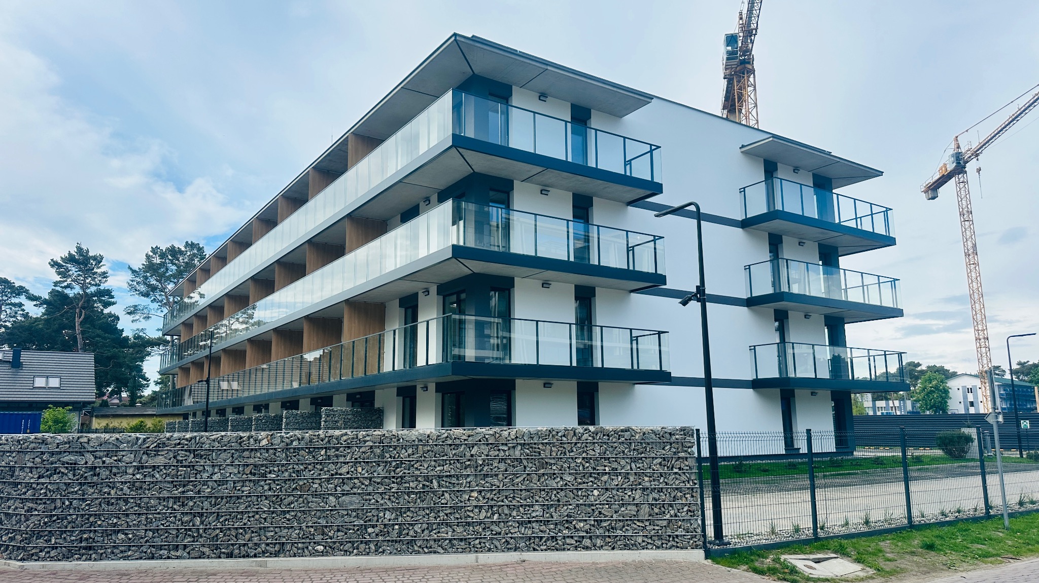 Nowoczesny, biały budynek wielorodzinny z przeszklonymi balkonami i gabionowym ogrodzeniem, w tle żurawie budowlane. Elewacja wykończona, stan deweloperski.