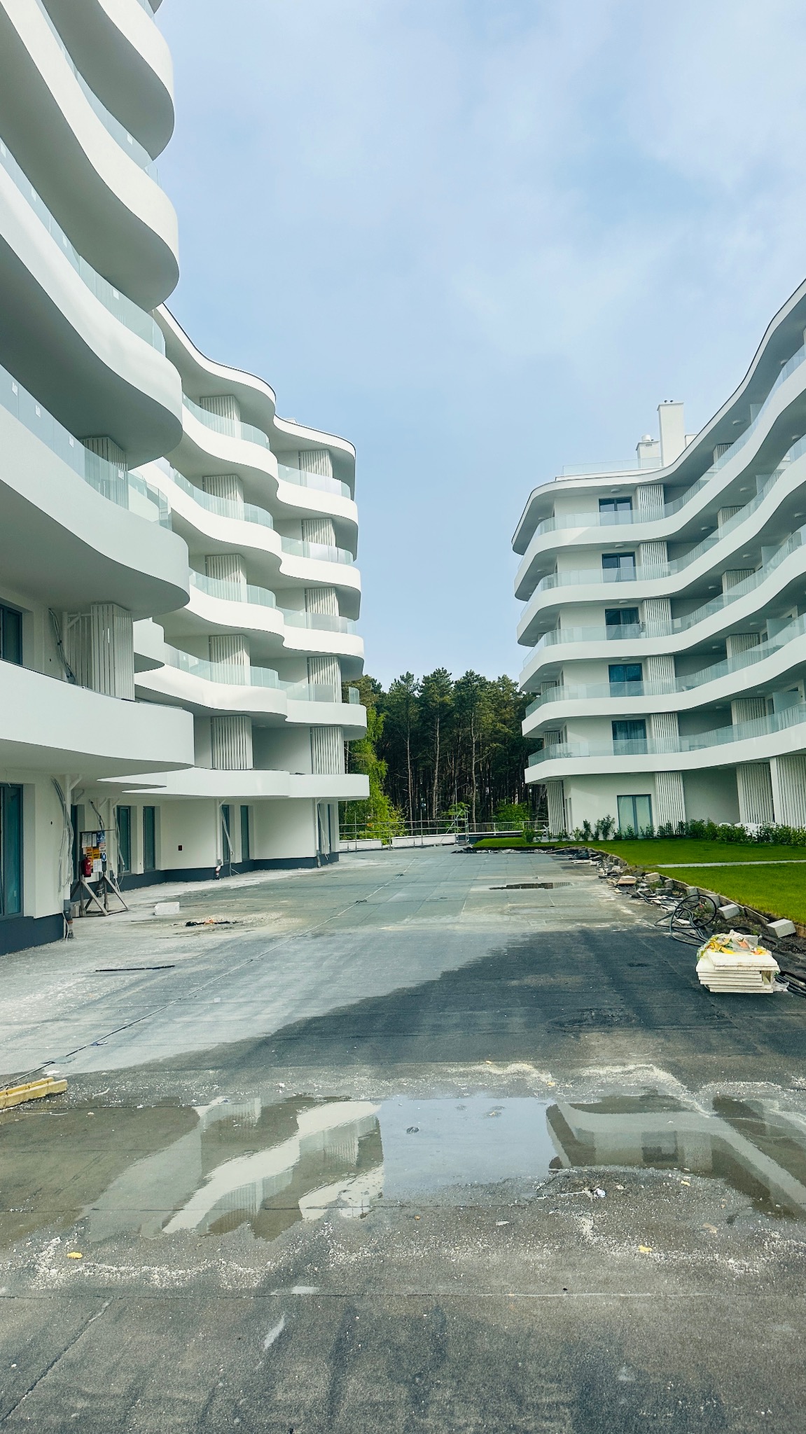 Nowoczesny budynek apartamentowy z białymi, falistymi balkonami i szklanymi balustradami. Widoczne prace wykończeniowe i odbicie budynków w kałużach.