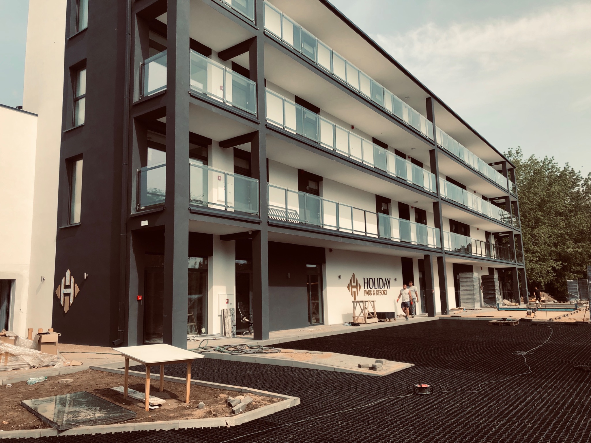 Nowoczesny budynek apartamentowy z balkonami wykończonymi szkłem, widoczne prace wykończeniowe terenu, w tle logo 'Holiday Park & Resort'.