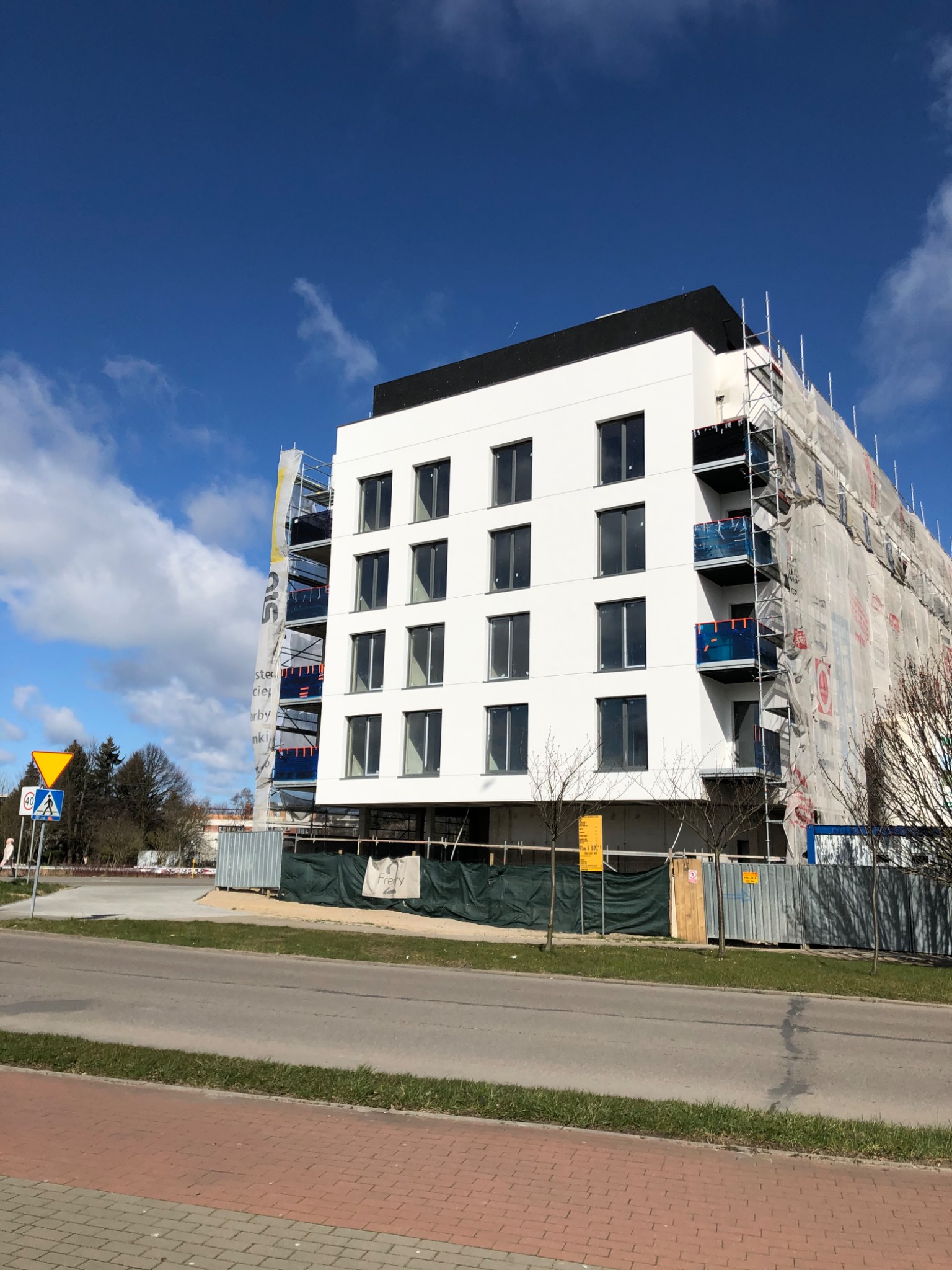 Nowoczesny budynek wielorodzinny w trakcie wykończenia elewacji, widoczne rusztowania i siatki ochronne, jasna fasada z ciemnymi oknami.