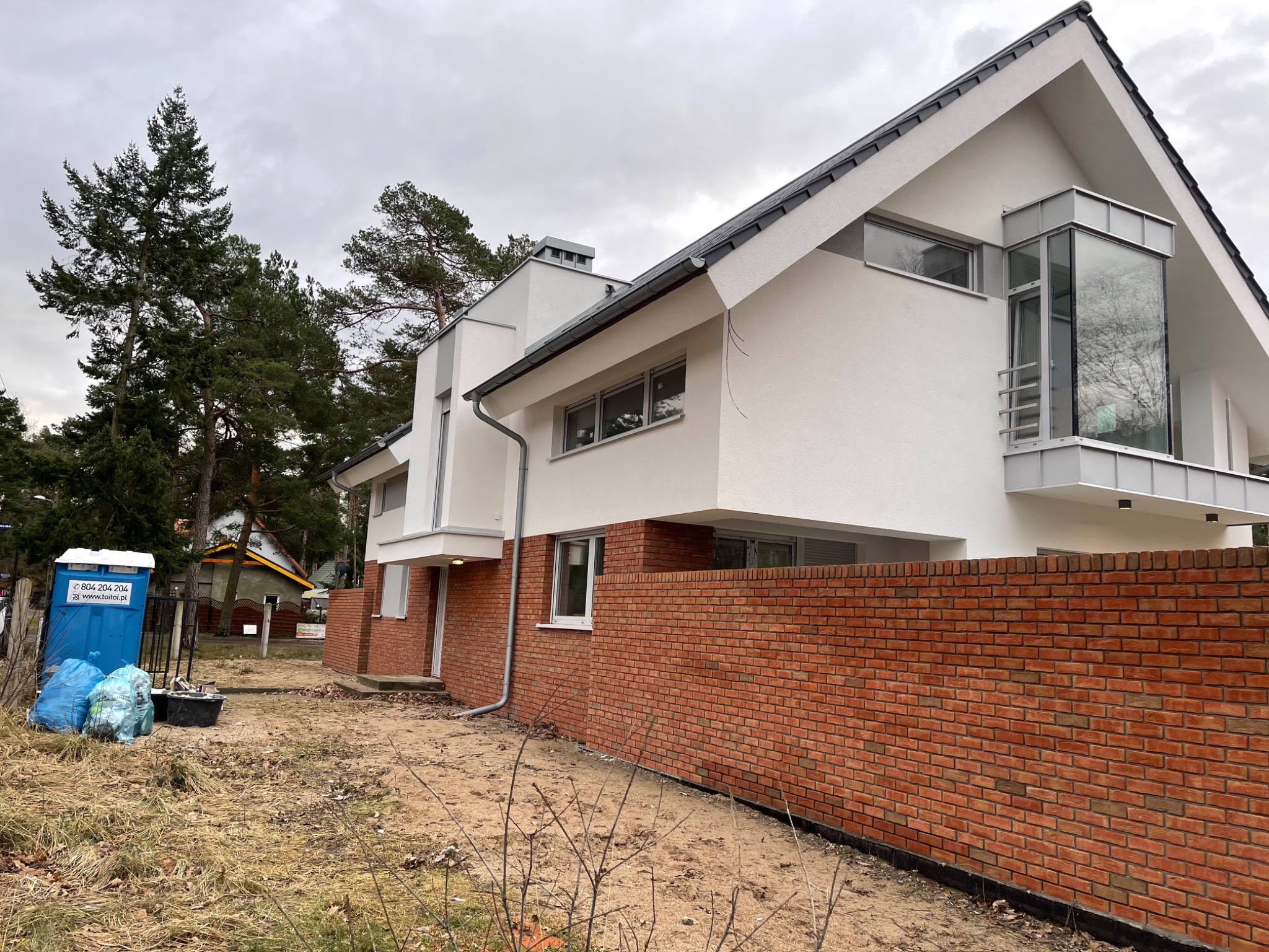 Nowoczesny dom jednorodzinny z białą elewacją i ceglanym murem oporowym, widok z zewnątrz na etapie wykończenia, z widoczną toaletą przenośną i workami na odpady budowlane.