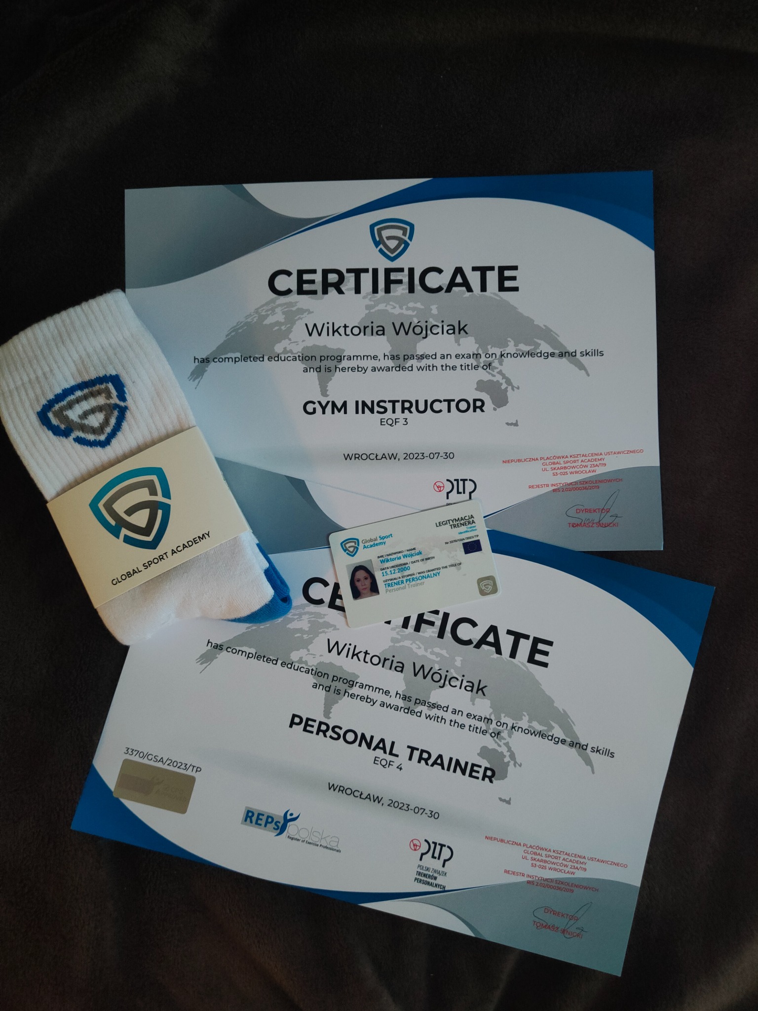 Dwa certyfikaty ukończenia kursów dla Wiktorii Wójciak: instruktora siłowni i trenera personalnego, legitymacja trenera personalnego z jej zdjęciem oraz skarpety z logo Global Sport Academy.