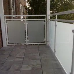 Balkon z białą, metalową balustradą i panelami z matowego szkła, widok na zewnątrz z szarymi płytkami podłogowymi.