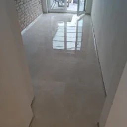 Świeżo ułożone, błyszczące płytki podłogowe w jasnym odcieniu beżu, z widocznym odbiciem okna tarasowego i ceglanej ściany, prowadzące na zewnątrz.