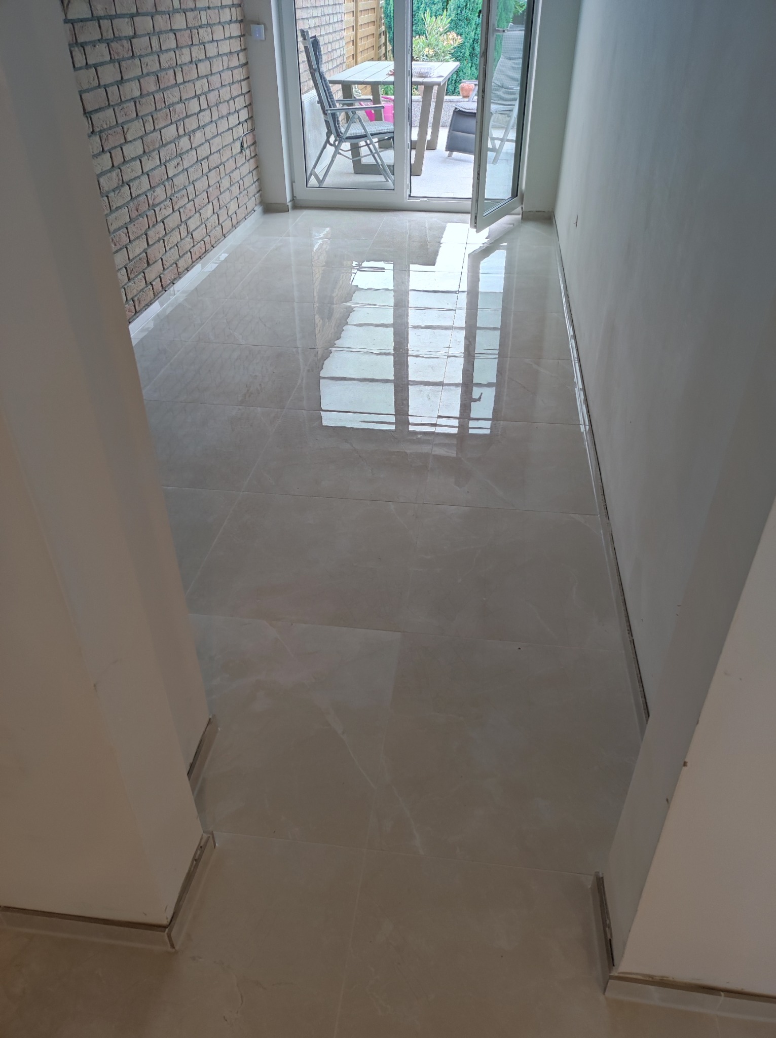 Świeżo ułożone, błyszczące płytki podłogowe w jasnym odcieniu beżu, z widocznym odbiciem okna tarasowego i ceglanej ściany, prowadzące na zewnątrz.