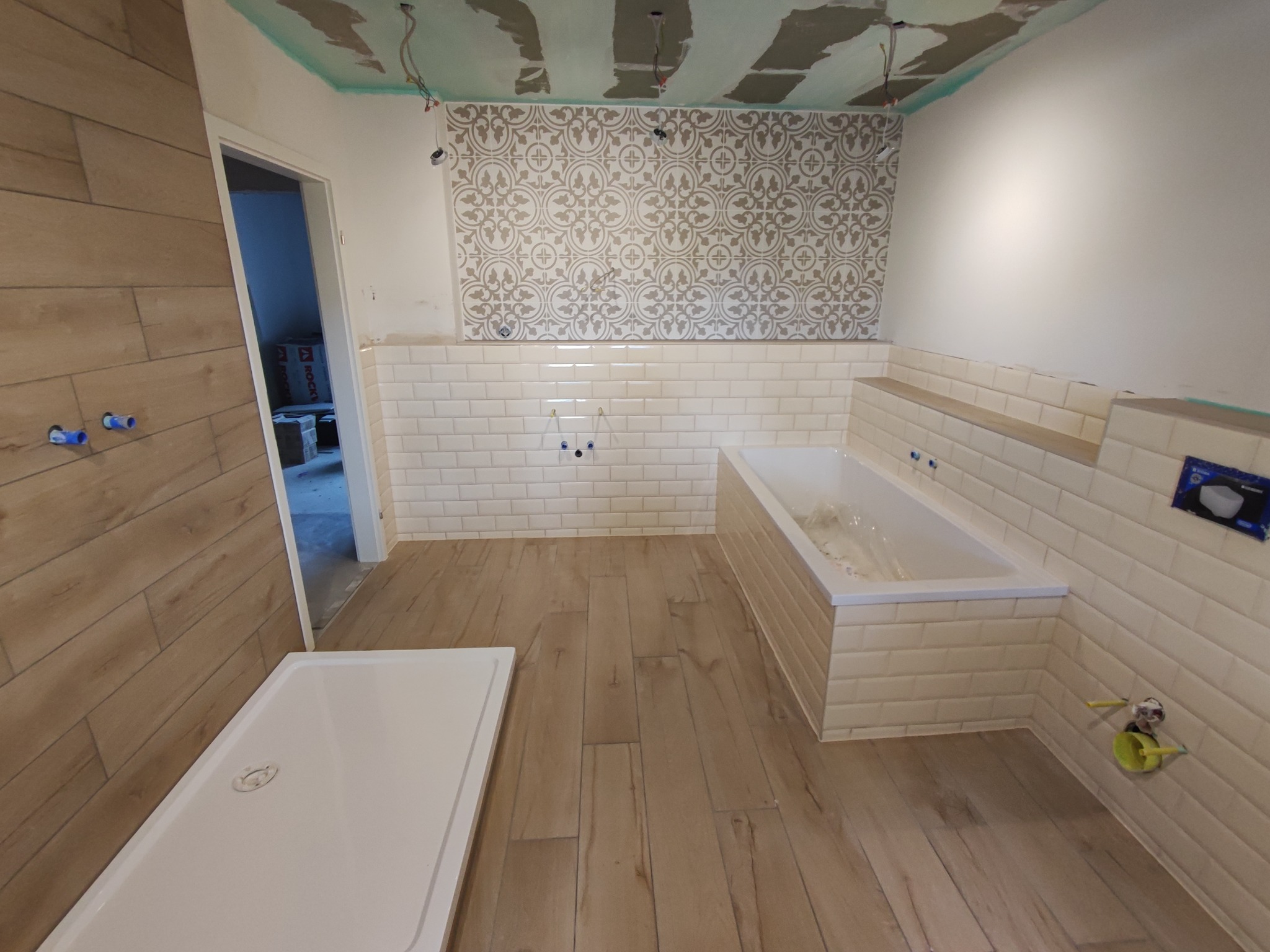 Łazienka w trakcie prac wykończeniowych: ułożone płytki typu metro na ścianach i obudowie wanny, wzorzysta tapeta na górnej części ściany, drewnopodobne panele na podłodze, widoczny brodzik...