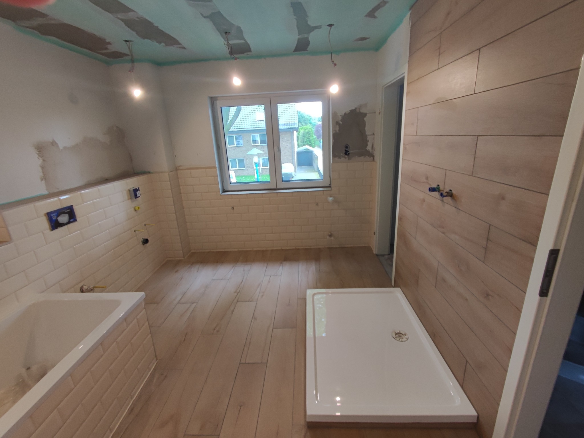 Niedokończona łazienka z białą wanną obudowaną płytkami typu metro, kabiną prysznicową z niskim brodzikiem, drewnopodobną podłogą i częściowo wykończonym sufitem z widocznymi przewodami.