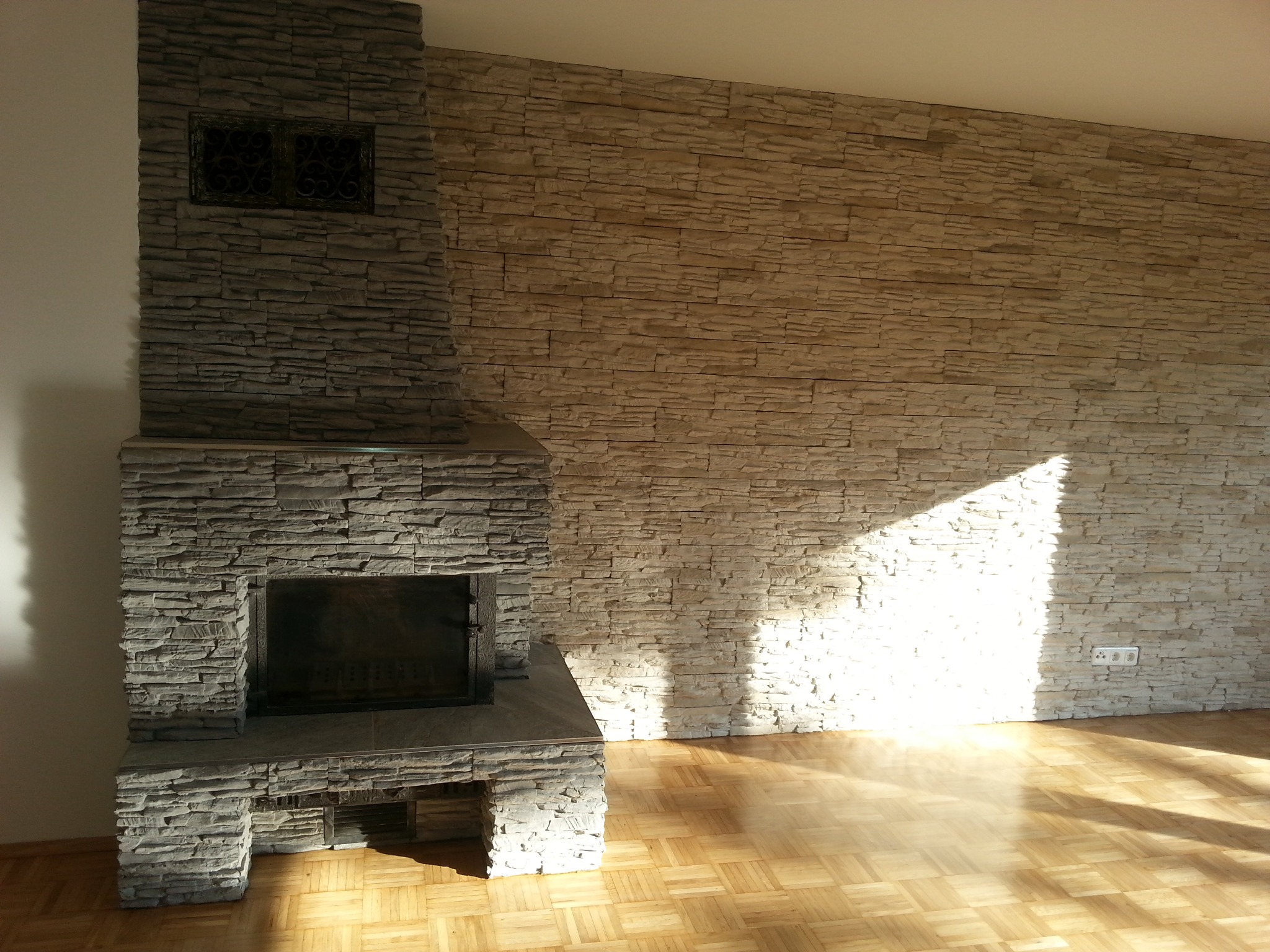 Wykończony kominek z szarego kamienia dekoracyjnego, sąsiadujący ze ścianą wyłożoną beżowym kamieniem, na parkiecie w jodełkę, oświetlony promieniami słońca.