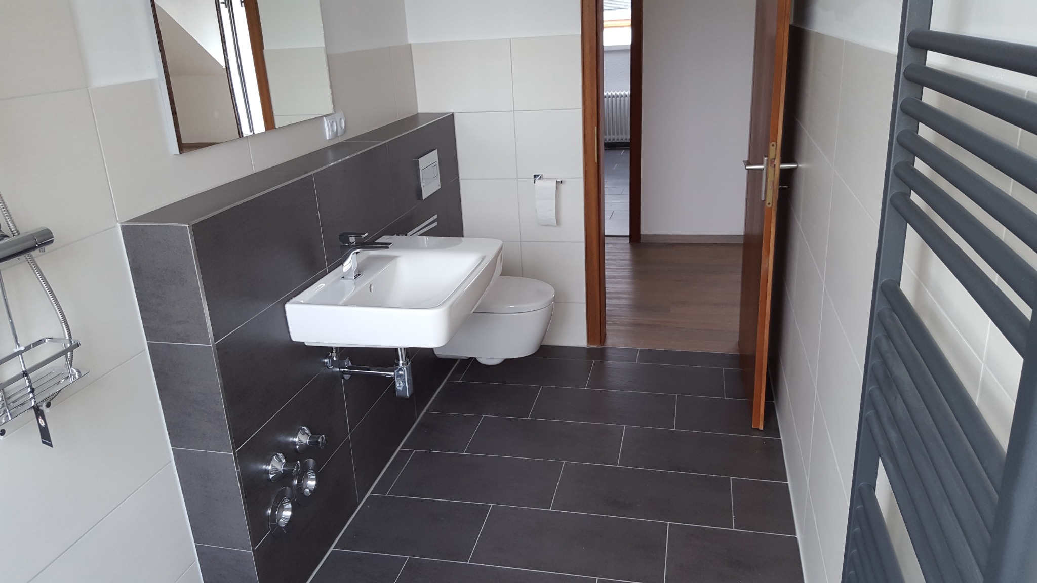 Nowoczesna łazienka z ciemnoszarą podłogą i ścianką pod umywalką, białą ceramiką sanitarną i grzejnikiem drabinkowym, widok na drewnianą podłogę w sąsiednim pomieszczeniu.