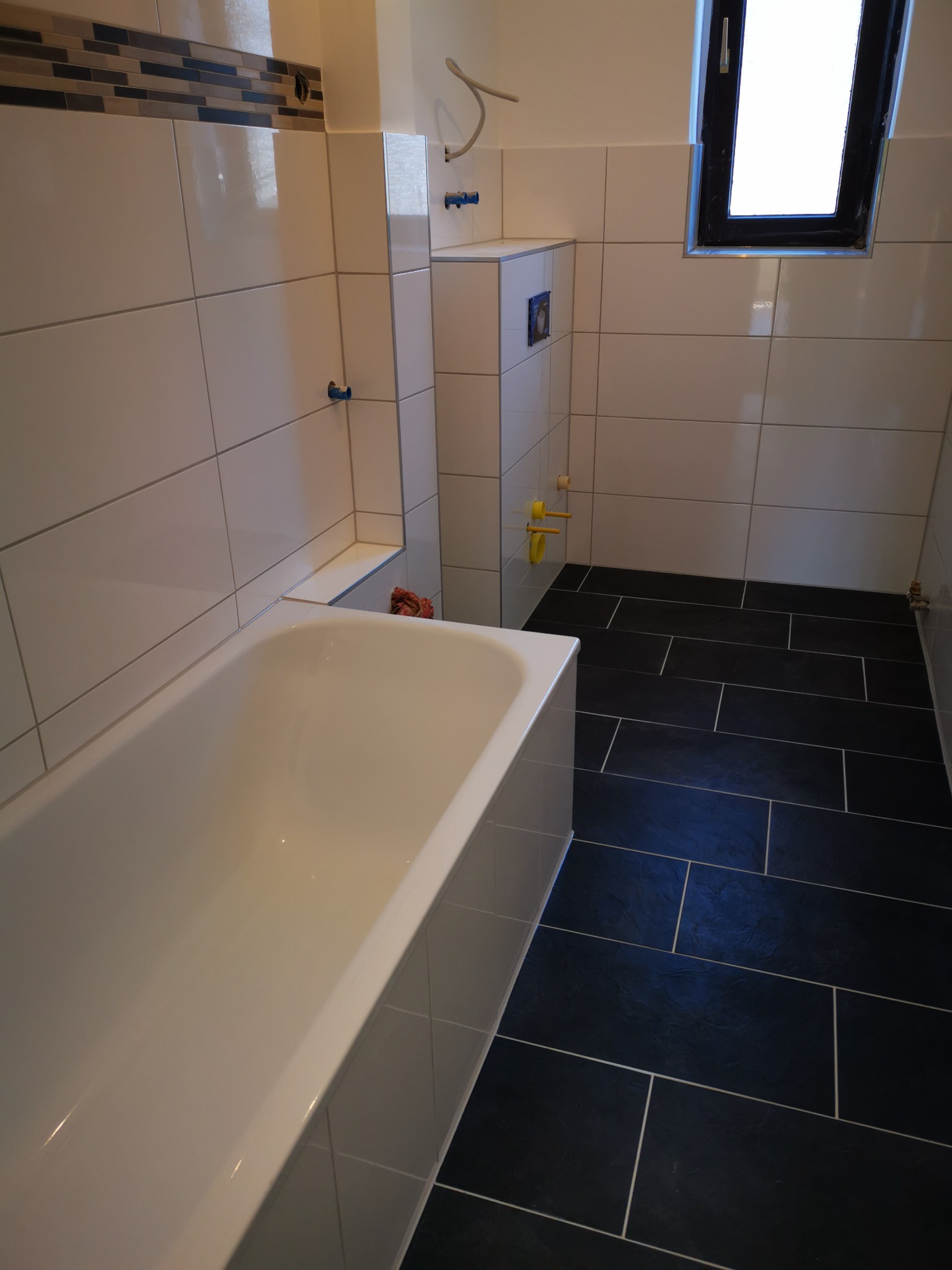 Wykończona łazienka z białą wanną, białymi płytkami ściennymi z dekoracyjnym paskiem i ciemnymi płytkami podłogowymi, widoczne instalacje hydrauliczne i okno.