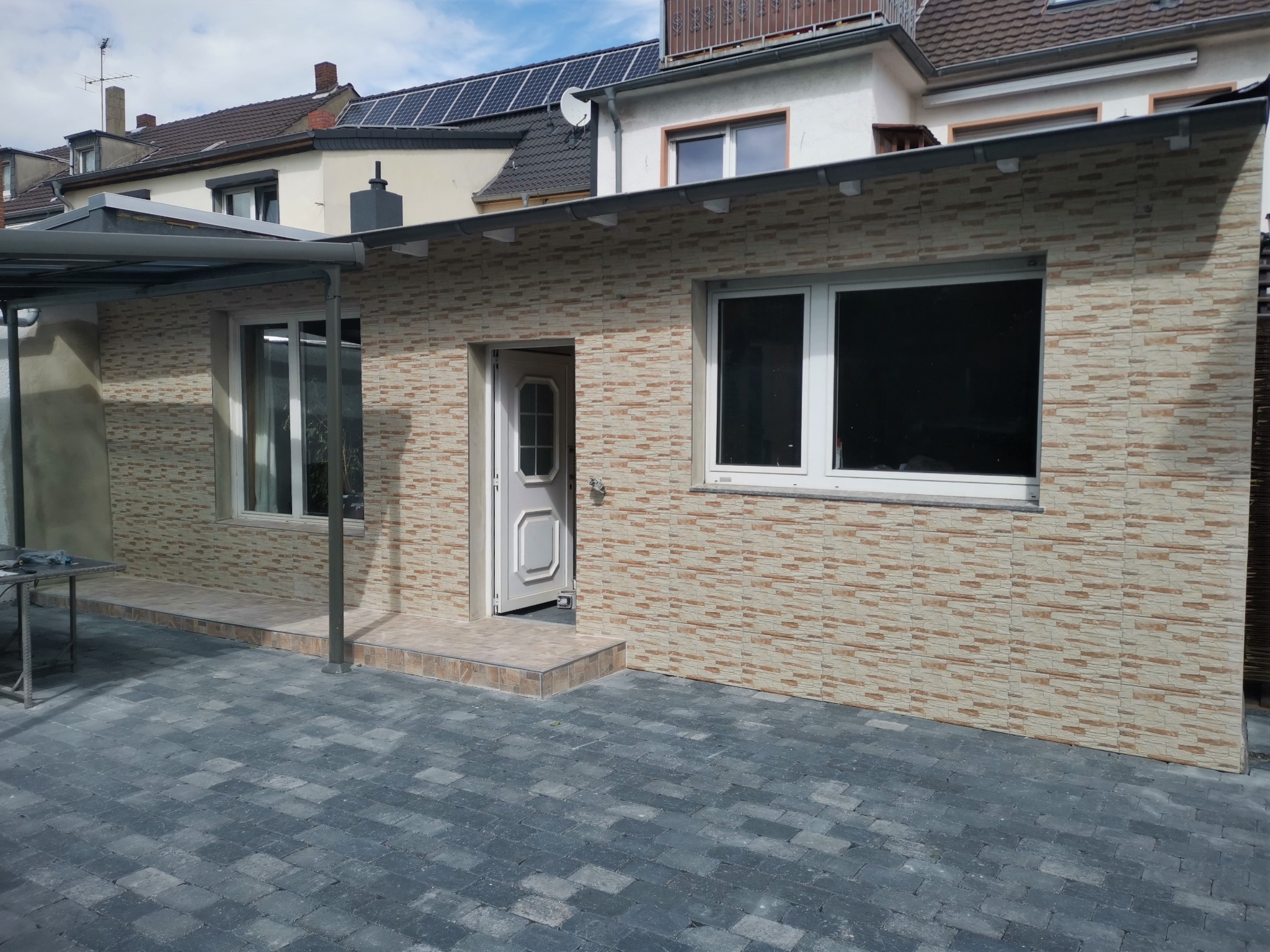 Elewacja budynku wykończona panelami imitującymi kamień, widoczne okna z białymi ramami, częściowo otwarte białe drzwi wejściowe, szara kostka brukowa przed budynkiem.