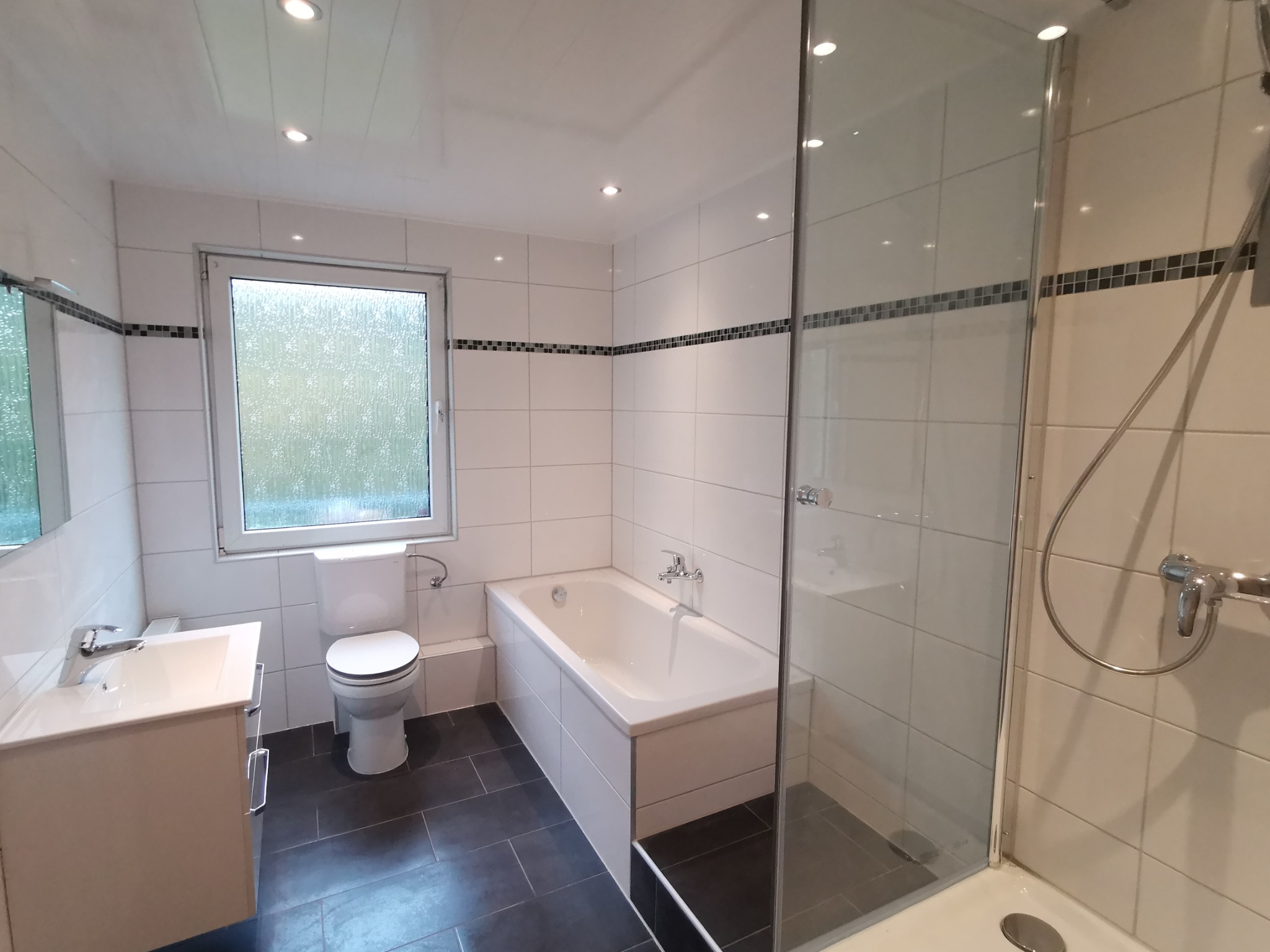 Jasna łazienka po remoncie z białymi płytkami, czarną podłogą, kabiną prysznicową ze szkła, umywalką z szafką i toaletą pod oknem.