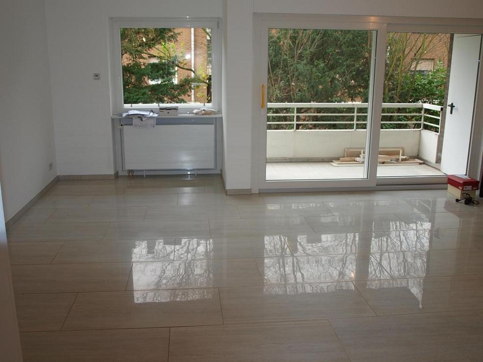 Jasne, błyszczące płytki podłogowe w pustym pokoju z wyjściem na balkon i oknem z widokiem na drzewa.