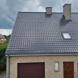 Nowy, błyszczący dach z ciemnej dachówki ceramicznej na domu z cegły, z dwoma kominami i oknem dachowym, na tle pochmurnego nieba.