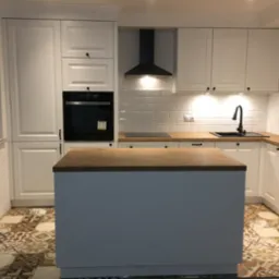 Biała kuchnia z wyspą i drewnianym blatem, wykończona płytkami typu metro, z dekoracyjną podłogą z płytek w stylu patchwork.