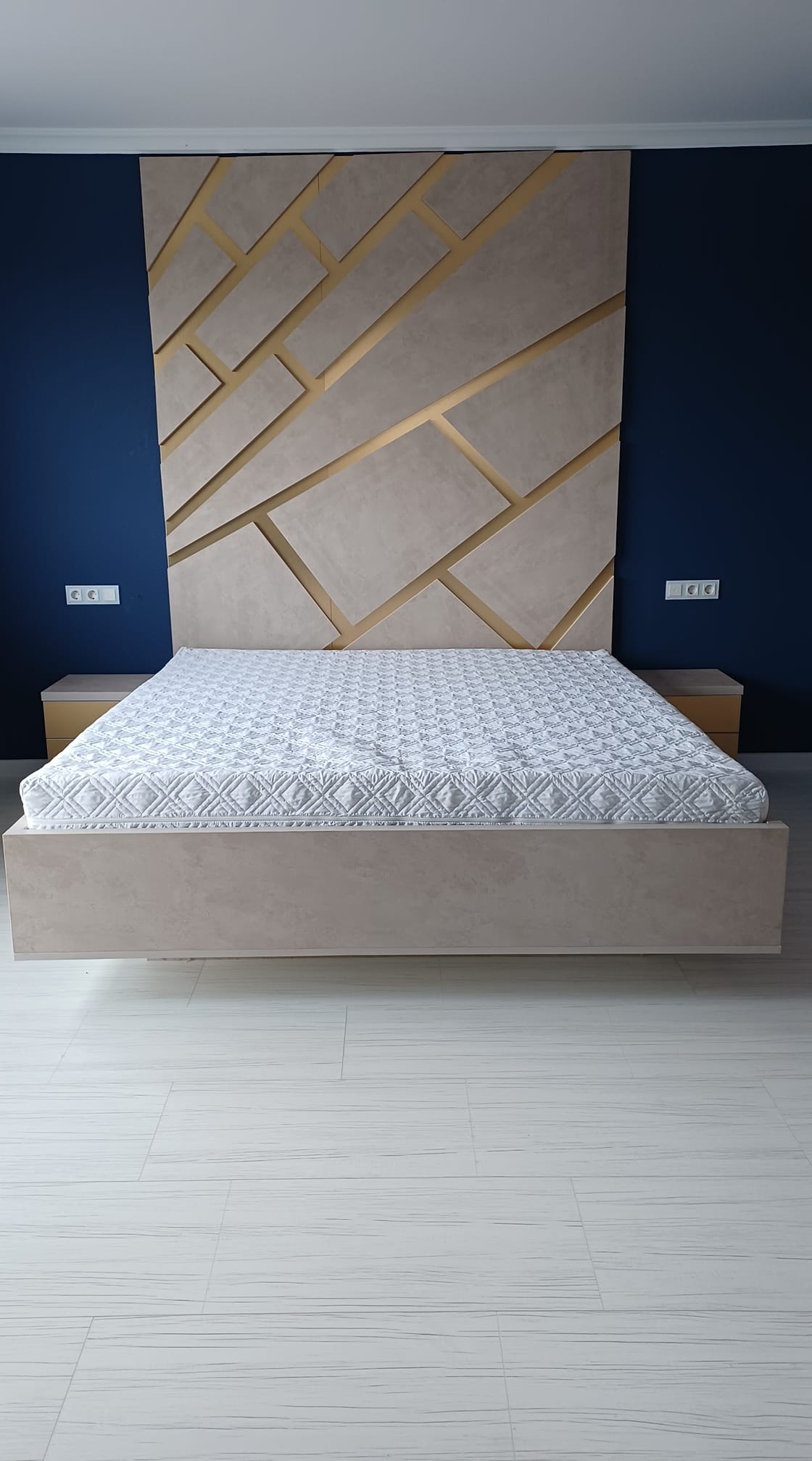 Nowoczesna sypialnia z łóżkiem na podeście i geometrycznym zagłówkiem w odcieniach beżu i złota na tle granatowej ściany, widoczne gniazdka elektryczne i biała podłoga.