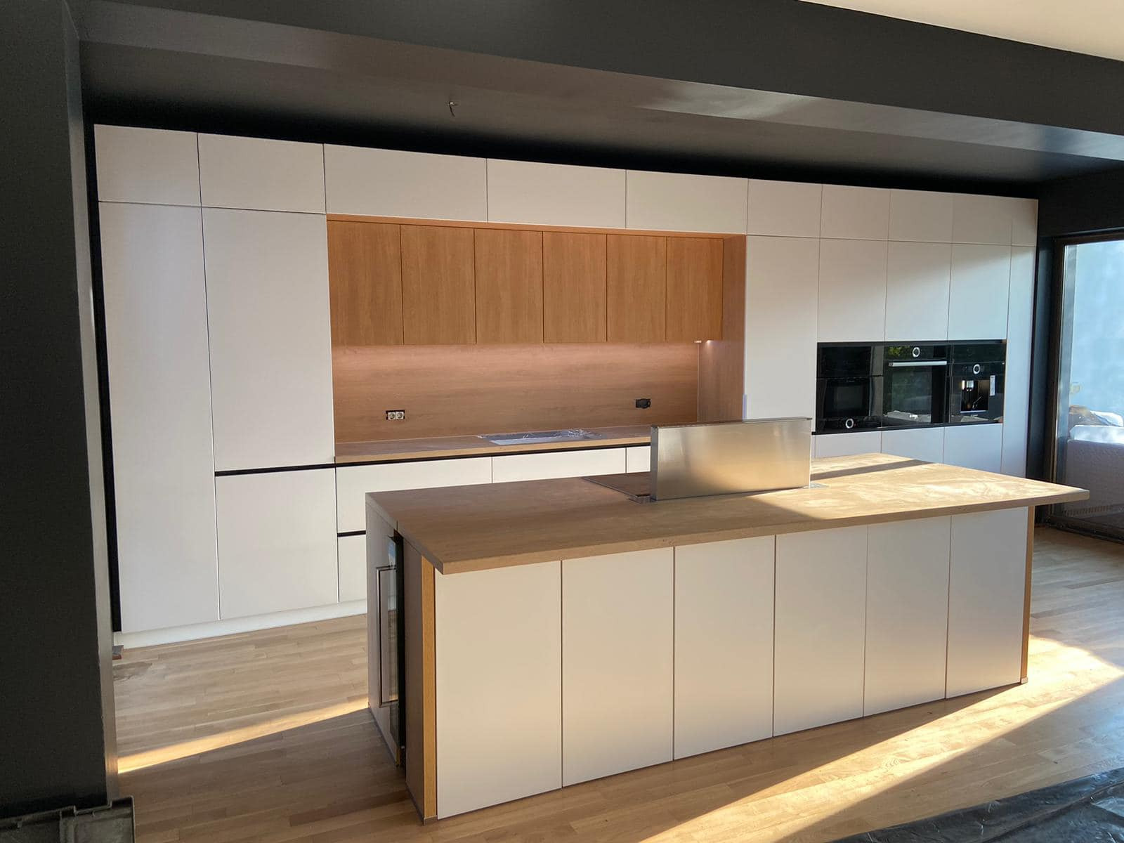 Nowoczesna kuchnia z białymi szafkami i drewnianymi akcentami, wyspa kuchenna z drewnianym blatem i wbudowaną chłodziarką na wino, oświetlenie LED pod szafkami wiszącymi, czarne elementy...