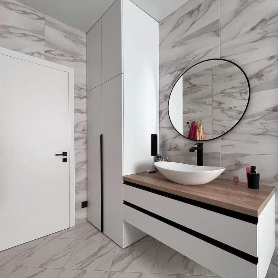 Nowoczesna łazienka z białą szafką pod umywalkę z drewnianym blatem, okrągłym lustrem i wysoką, białą szafą w zabudowie, marmurowe płytki na ścianach i podłodze.