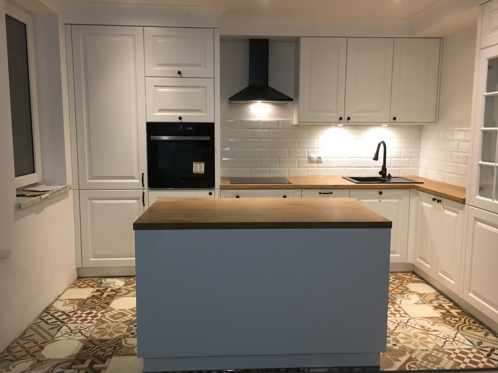 Biała kuchnia z wyspą i drewnianym blatem, wykończona płytkami typu metro, z dekoracyjną podłogą z płytek w stylu patchwork.