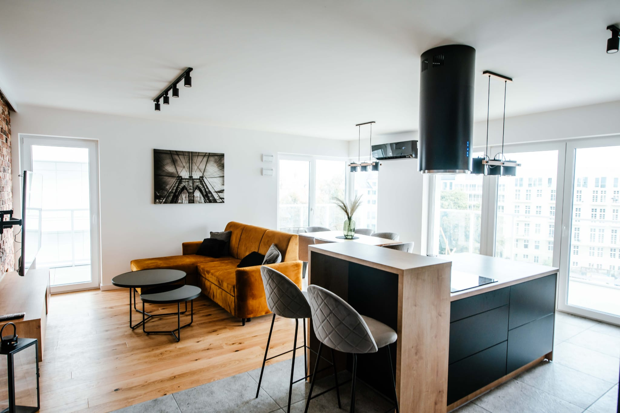 Nowoczesne wnętrze salonu z aneksem kuchennym w Kętach: drewniana podłoga, musztardowa sofa, czarne stoliki kawowe i wyspa kuchenna z hokerami.