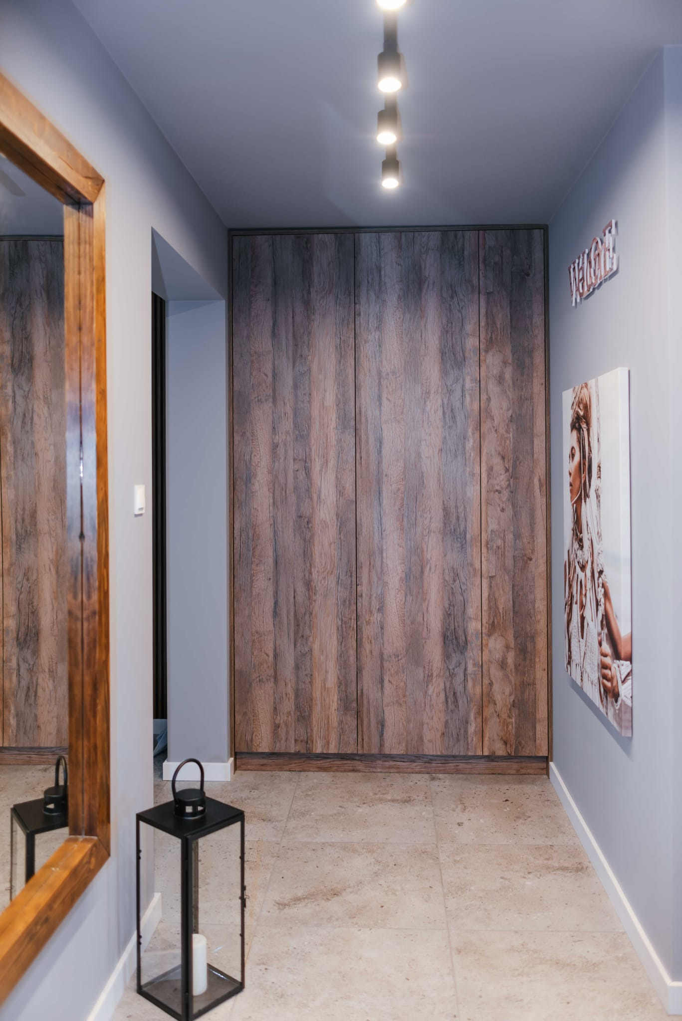 Korytarz z dużą, drewnianą szafą wnękową o fakturze naturalnego drewna, lustro w drewnianej ramie, dekoracje ścienne i podłoga wyłożona jasnymi płytkami.