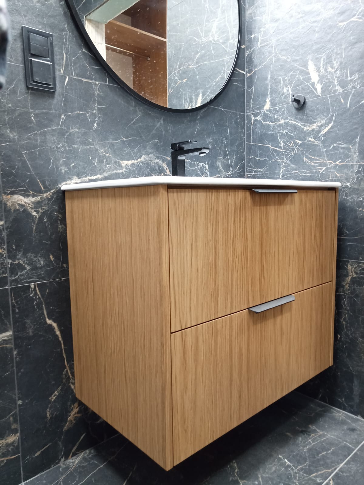 Nowoczesna łazienka z wiszącą szafką pod umywalkę z drewnianym frontem, czarną armaturą i lustrem w okrągłej ramie na tle ściany wyłożonej płytkami imitującymi czarny marmur.