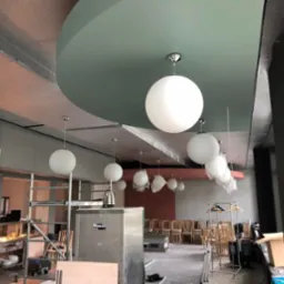 Wnętrze lokalu gastronomicznego w trakcie wykańczania, widoczne wiszące, kuliste lampy, częściowo jeszcze w folii ochronnej, surowy sufit z widoczną instalacją, krzesła ustawione w rzędach...