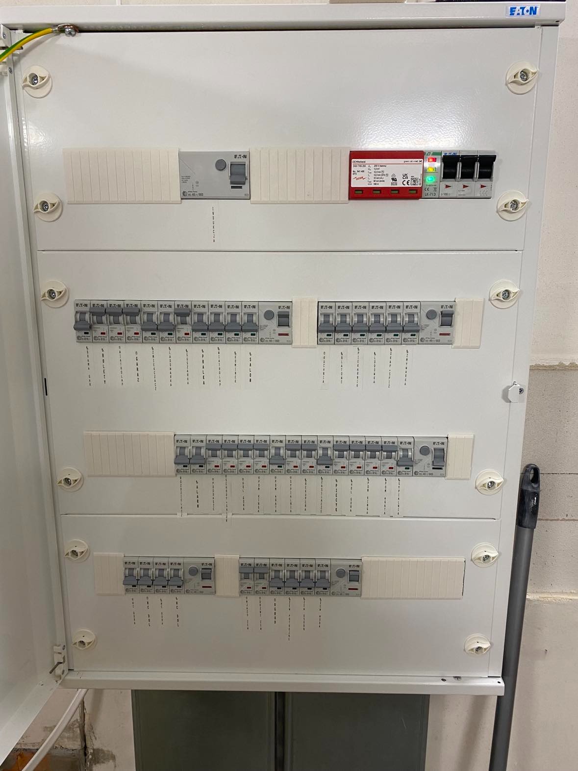 Otwarta, biała skrzynka elektryczna z zainstalowanymi bezpiecznikami i wyłącznikami różnicowoprądowymi marki EATON; widoczne oznaczenia obwodów na naklejkach pod wyłącznikami.